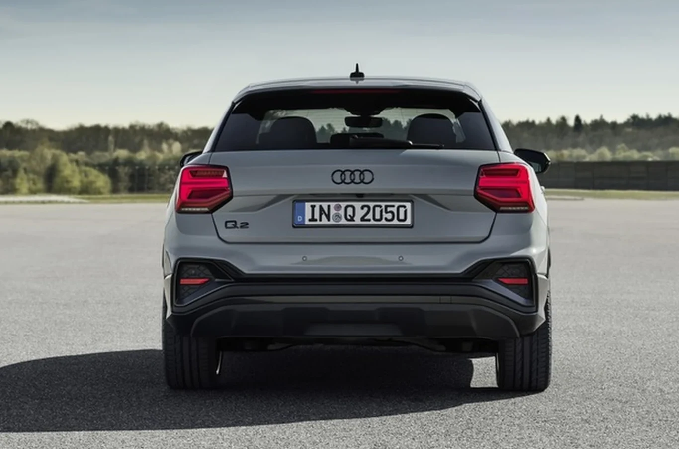 Bagażnik o pojemności 405-1050 litrów (50 litrów mniej w wersjach z napędem quattro) nie robi wrażenia na tle konkurencji, ale jest praktycznie wykończony i łatwy w załadunku. szary samochód Audi Q2 stoi na asfalcie, tylne światła są włączone, w tle widoczne zielone drzewa i niebo