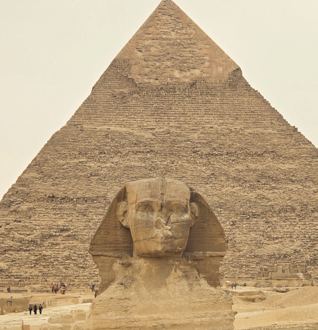 Wielki Sfinks w kompleksie piramid w Gizie. Wielki Sfinks z masywnym tułowiem i ludzką głową umieszczony na tle ogromnej piramidy Cheopsa, otoczony piaskami pustyni i niewielkimi sylwetkami ludzi w oddali.