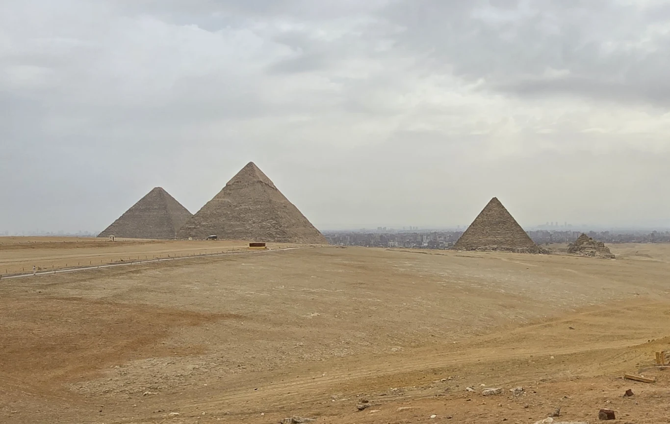 Piramidy w Gizie Trzy wielkie piramidy w Gizie na pustynnym terenie, w tle miasto i szare, zachmurzone niebo.
