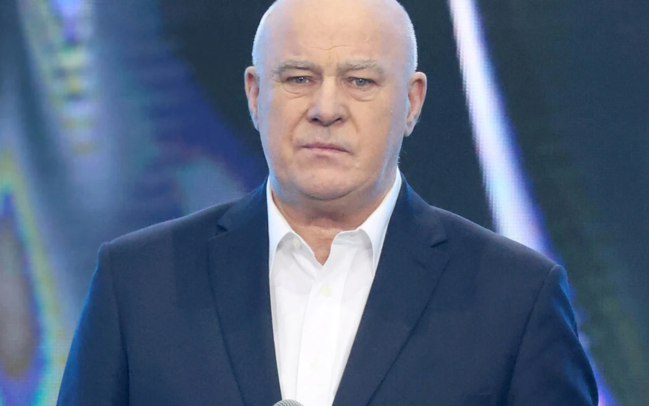 Ryszard Rynkowski Ryszard Rynkowski na scenie w ciemnym garniturze i białej koszuli, z mikrofonem, na tle niebiesko-fioletowym.