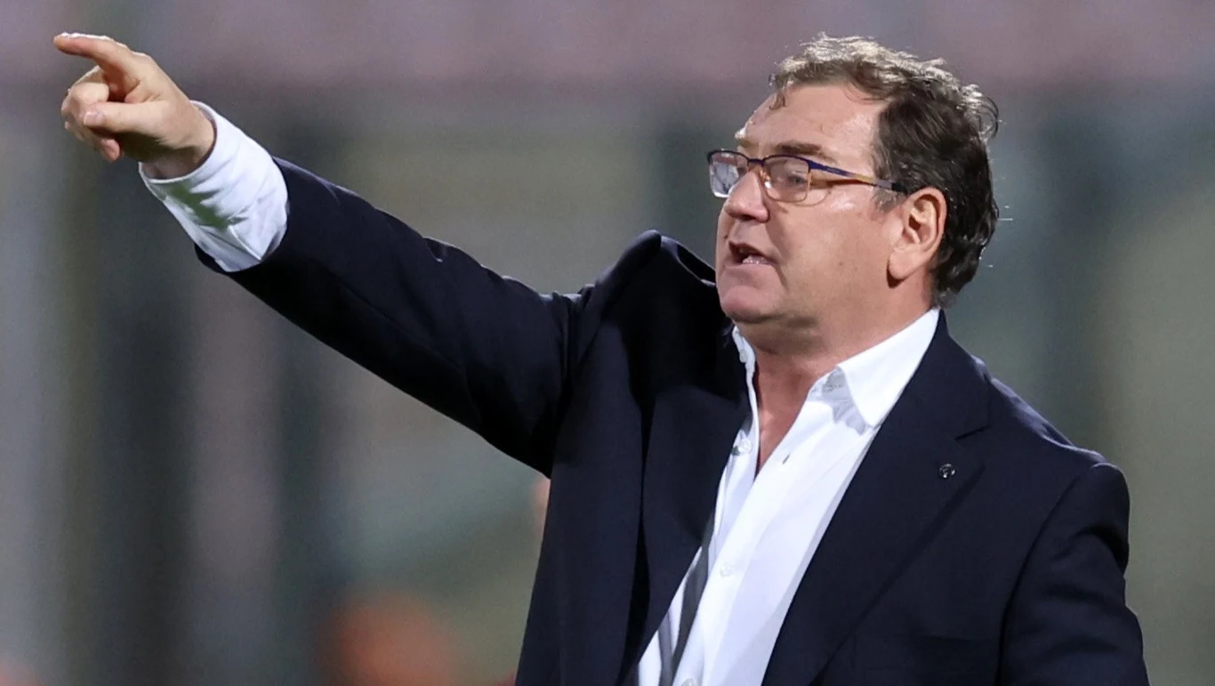 mężczyzna w okularach ubrany w ciemny garnitur i białą koszulę energicznie wskazuje ręką w bok, ma poważny wyraz twarzy