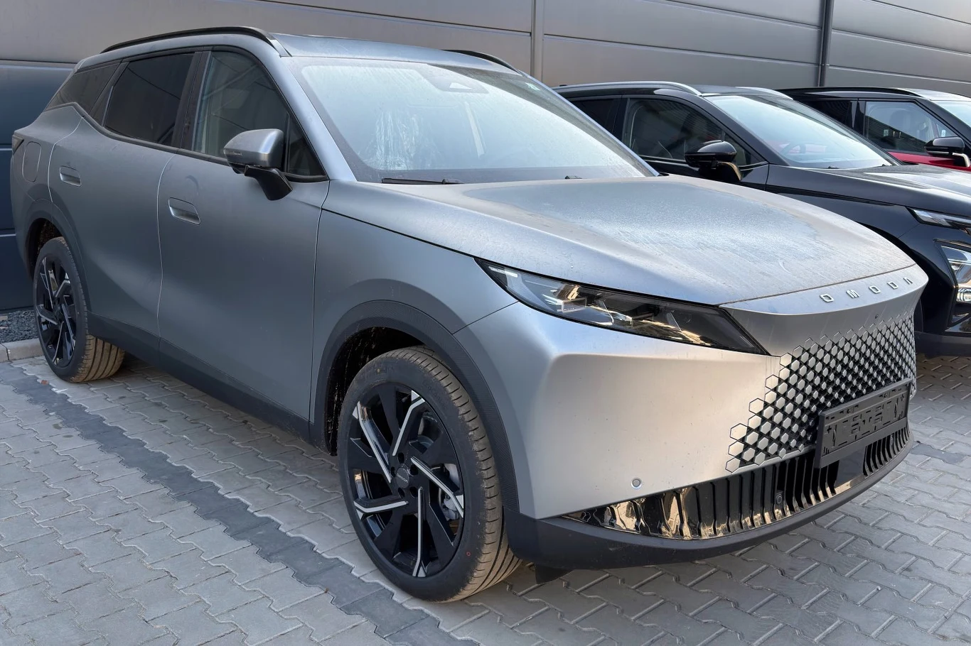 srebrnoszary SUV o nowoczesnej sylwetce i eleganckiej, oszczędnej stylistyce, wyróżniający się dużym, charakterystycznym grillem z geometrycznym wzorem oraz ostro zarysowanymi reflektorami, zaparkowany na kostce brukowej obok innych samochodów