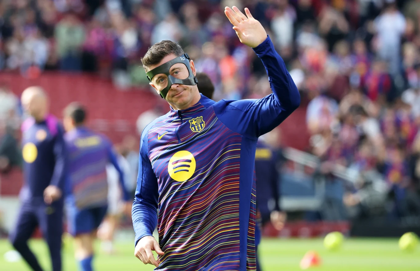 Robert Lewandowski w stroju treningowym Barcelony, z czarną maską na twarzy, unosi dłoń na stadionie.