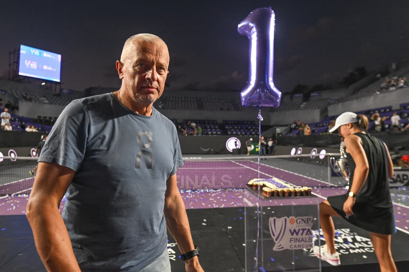 mężczyzna w szarej koszulce stoi na korcie tenisowym, w tle balon w kształcie cyfry 1 oraz dekoracje związane z wydarzeniem sportowym, po prawej stronie osoba z trofeum