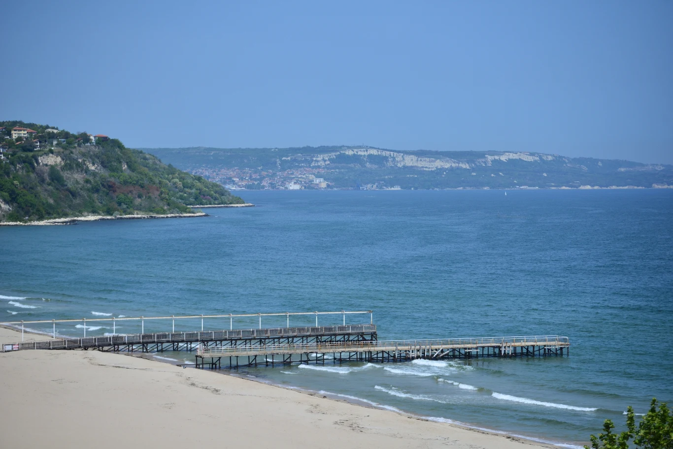 drewniane molo wychodzące w niebieskie morze, szeroka, pusta plaża oraz zalesione wzgórza w tle