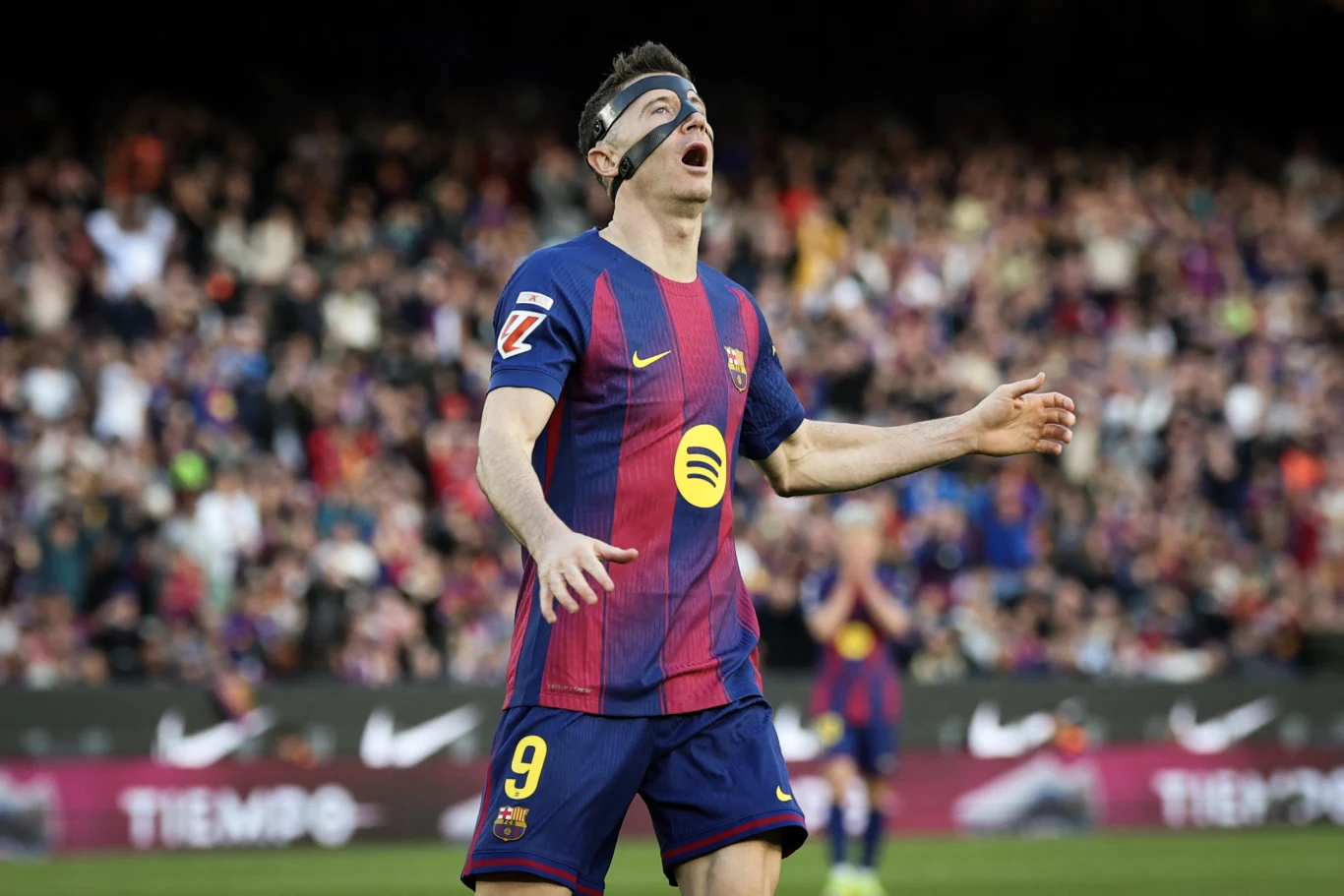 Piłkarz FC Barcelony w granatowo-bordowej koszulce z numerem 9 i czarną opaską na twarzy gestykuluje na tle tłumu kibiców na stadionie.