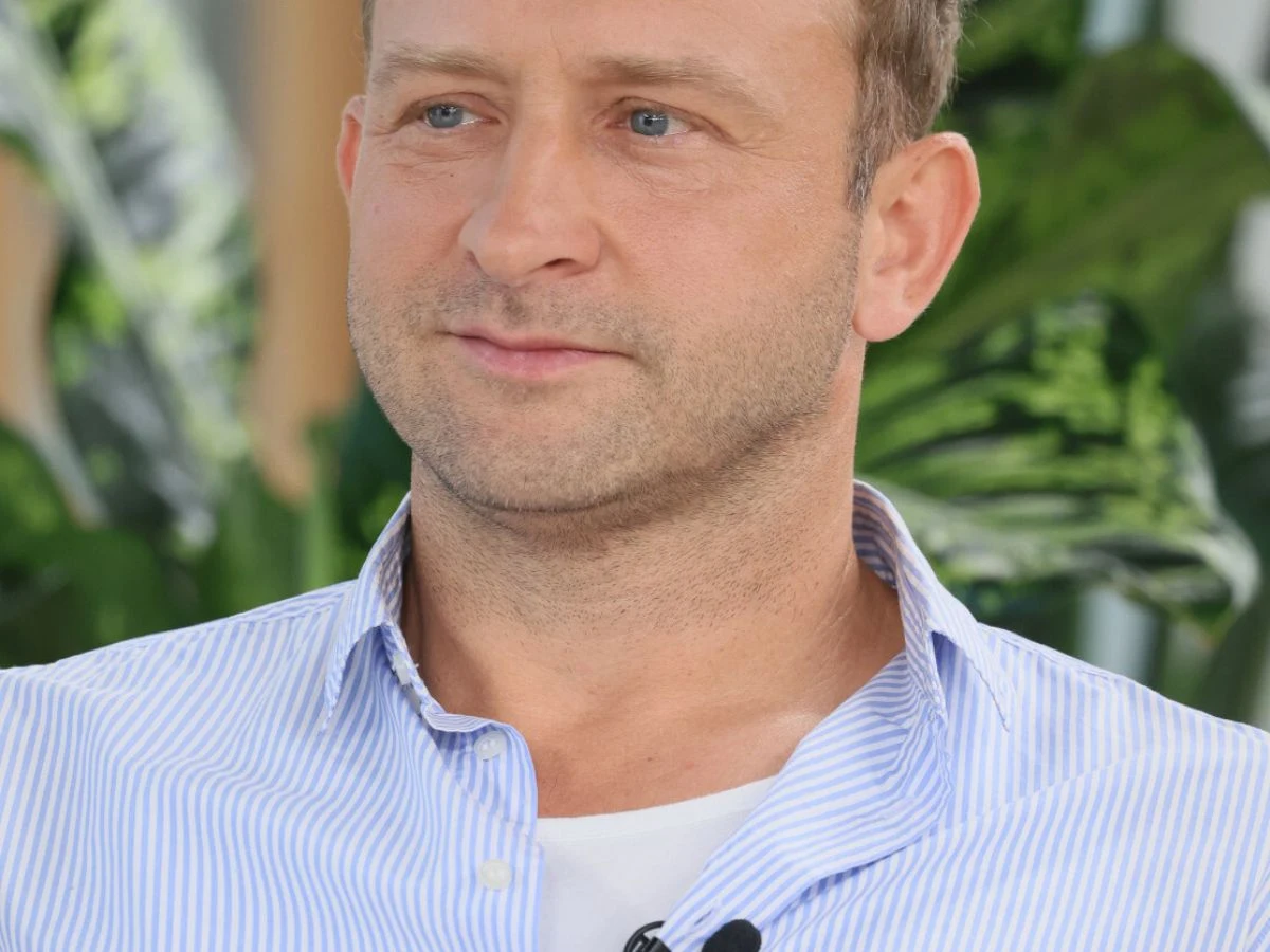 Mężczyzna w jasnoniebieskiej koszuli w paski z białym podkoszulkiem, siedzi na tle zielonych roślin, na koszuli przypięty mikrofon.