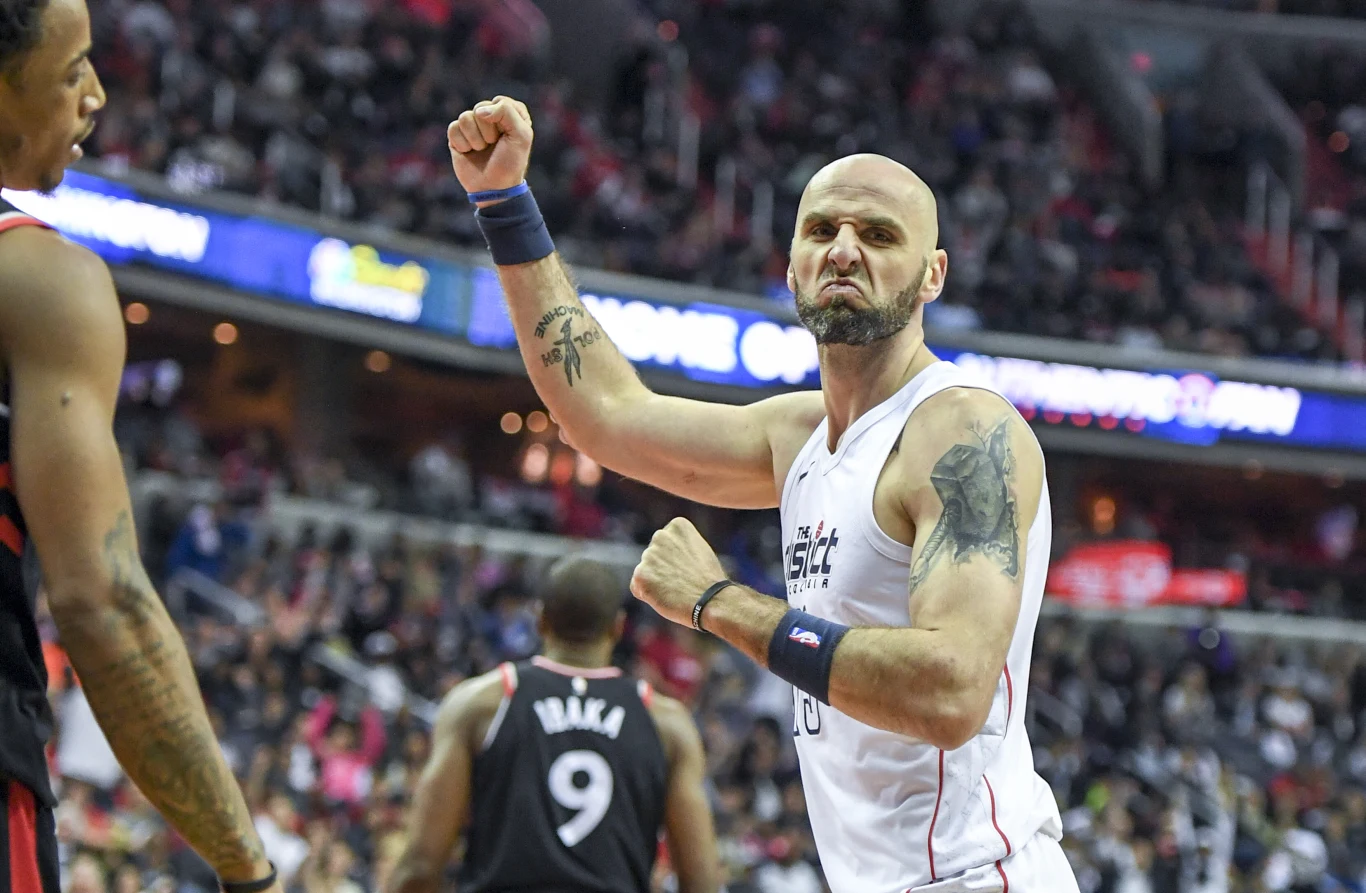 koszykarz w białym stroju z zaciśniętą pięścią oraz wyraźną mimiką twarzy wyrażającą intensywne emocje, w tle zawodnicy rywali w czarnych koszulkach oraz rozmyta publiczność na trybunach dużej hali sportowej