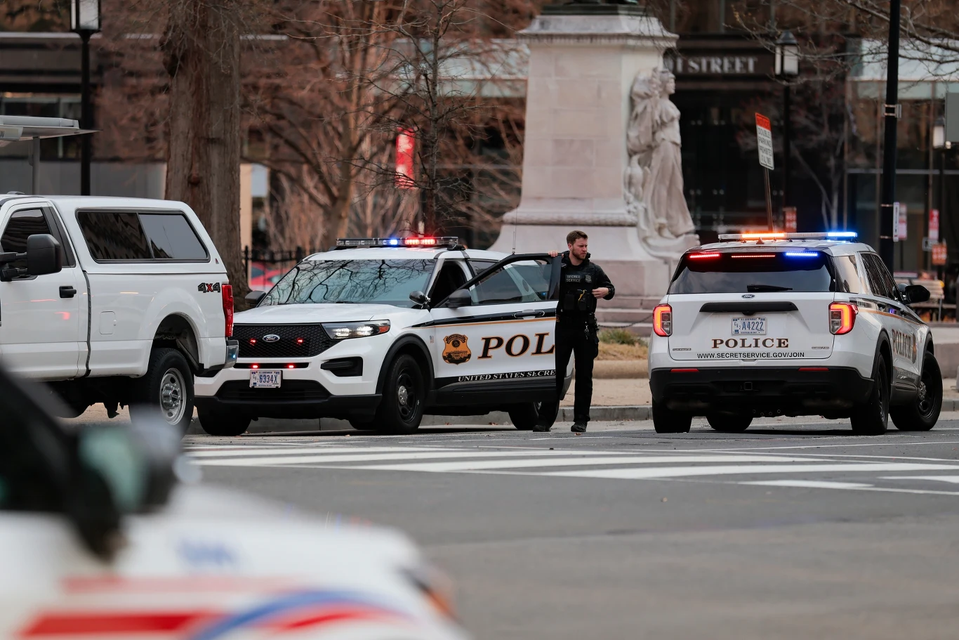 Dwóch radiowozów policyjnych z włączonymi światłami ostrzegawczymi zatrzymanych na skrzyżowaniu, policjant stoi między pojazdami, w tle pomnik oraz drzewa bez liści.
