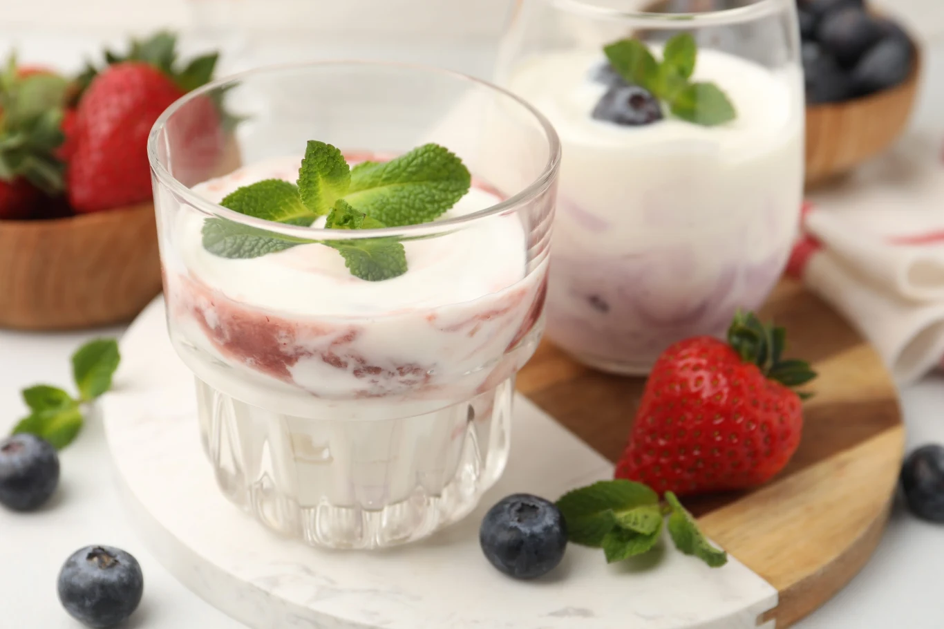 Dwie szklanki jogurtu z warstwą owoców, udekorowane świeżą miętą, otoczone truskawkami i borówkami rozmieszczonymi na marmurowej desce.