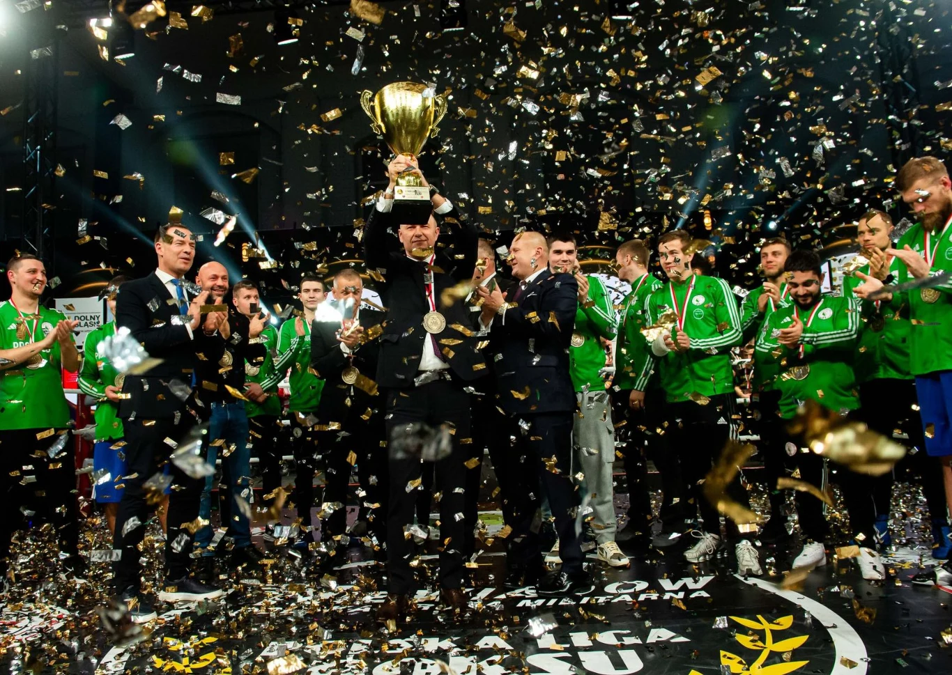 zespół ubrany w zielone stroje sportowe świętuje zwycięstwo, jeden z mężczyzn trzyma dużego, złotego pucharu nad głową, wokół spadają złote konfetti, a inni uczestnicy biją brawo i wyrażają radość