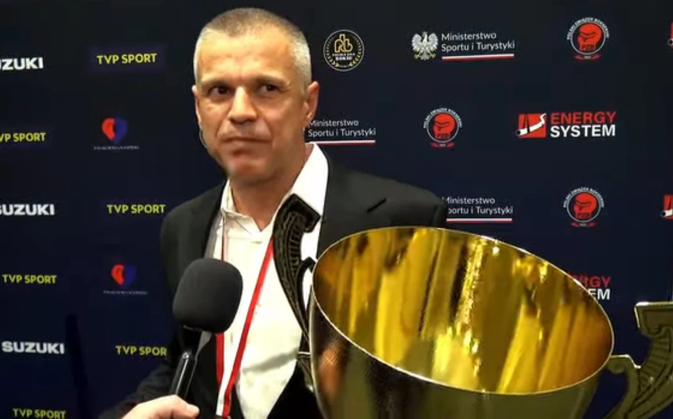 Mężczyzna w garniturze trzyma dużego, złotego pucharu, a obok niego znajduje się mikrofon skierowany w jego stronę; tło stanowi ściana z logotypami sponsorów i Ministerstwa Sportu i Turystyki.