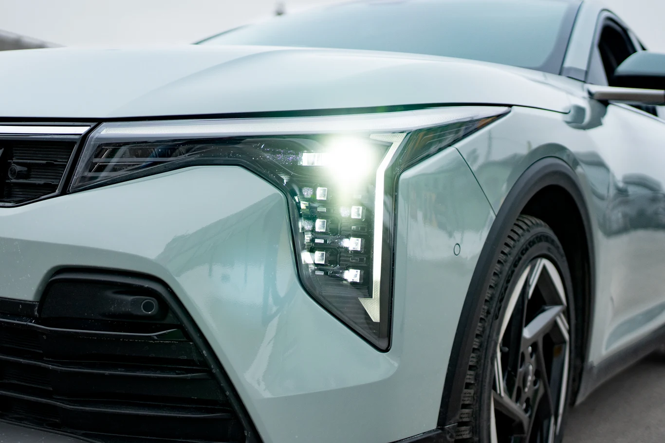 nowoczesny samochód w jasnym kolorze, z włączonymi reflektorami LED o charakterystycznym, geometrycznym kształcie, widoczne ostre linie maski i felga o oryginalnym wzorze