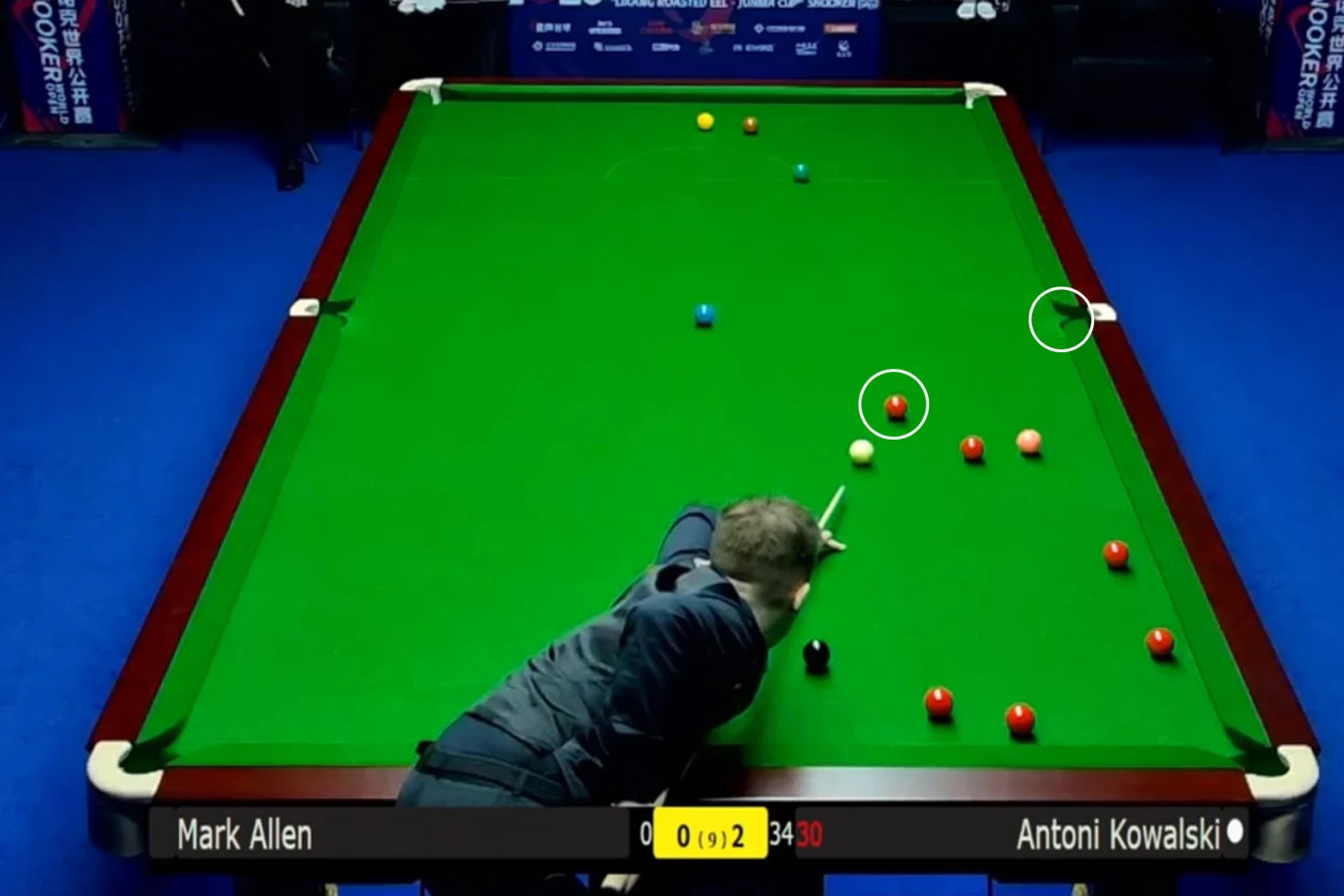 zawodnik wykonuje uderzenie kijem bilardowym na stole snookerowym, bilki ustawione są w różnych miejscach na zielonym suknie, na planszy wyników widnieją nazwiska Mark Allen i Antoni Kowalski oraz aktualny wynik meczu