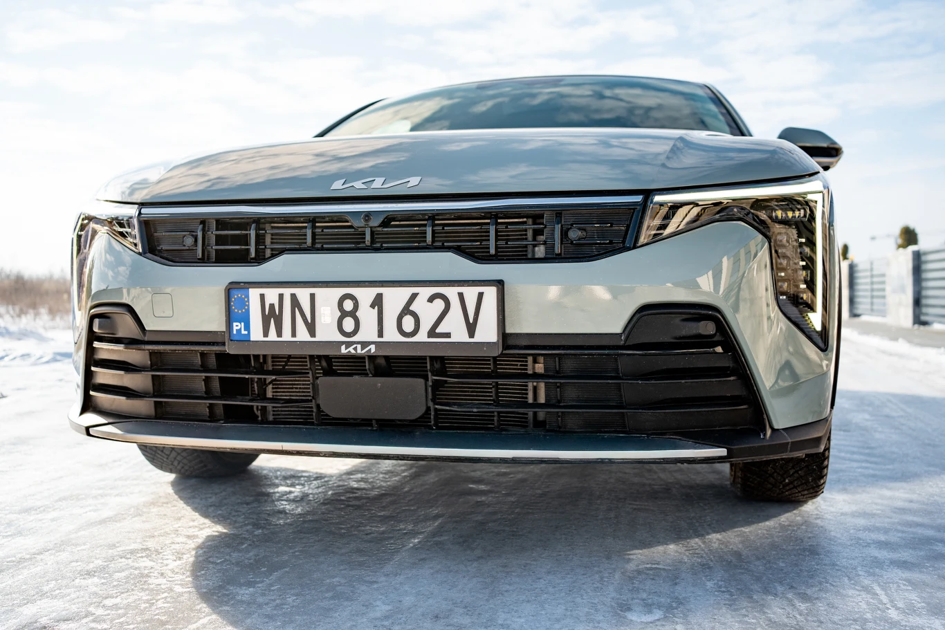 Przód srebrnego samochodu marki Kia z wyraźnie widoczną nowoczesną osłoną chłodnicy, charakterystycznymi reflektorami LED oraz polską tablicą rejestracyjną na tle zimowej scenerii.