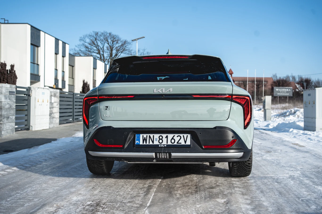 tył jasnego nowoczesnego samochodu Kia z wyraźnym oznaczeniem marki i rejestracją, na zaśnieżonej drodze pomiędzy nowoczesnymi budynkami mieszkalnymi