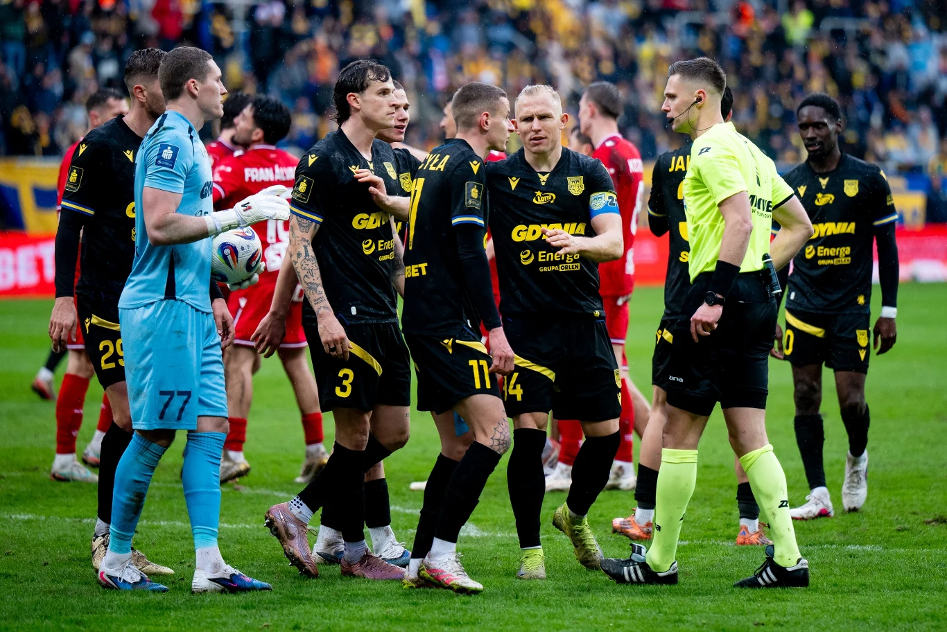 grupa piłkarzy w czarnych strojach gromadzi się wokół sędziego w żółtym stroju podczas intensywnej dyskusji na boisku, w tle widoczni zawodnicy drużyny przeciwnej w czerwonych koszulkach oraz kibice na trybunach