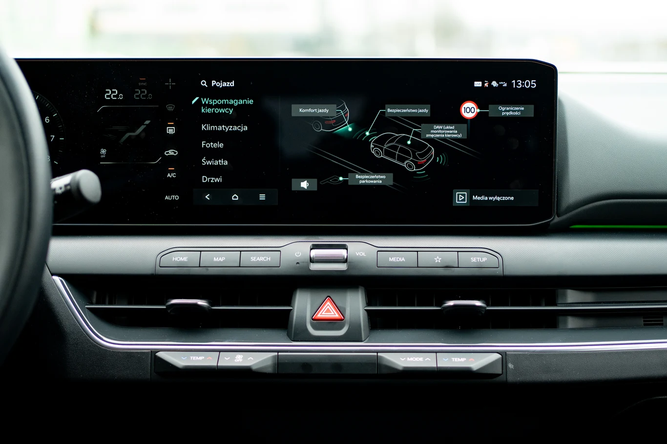Dotykowy ekran systemu multimedialnego samochodu wyświetlający menu ustawień pojazdu, funkcje wspomagania kierowcy oraz informacje o zużyciu energii, poniżej centralnie umieszczony przycisk świateł awaryjnych i panel przycisków obsługujących system aud...