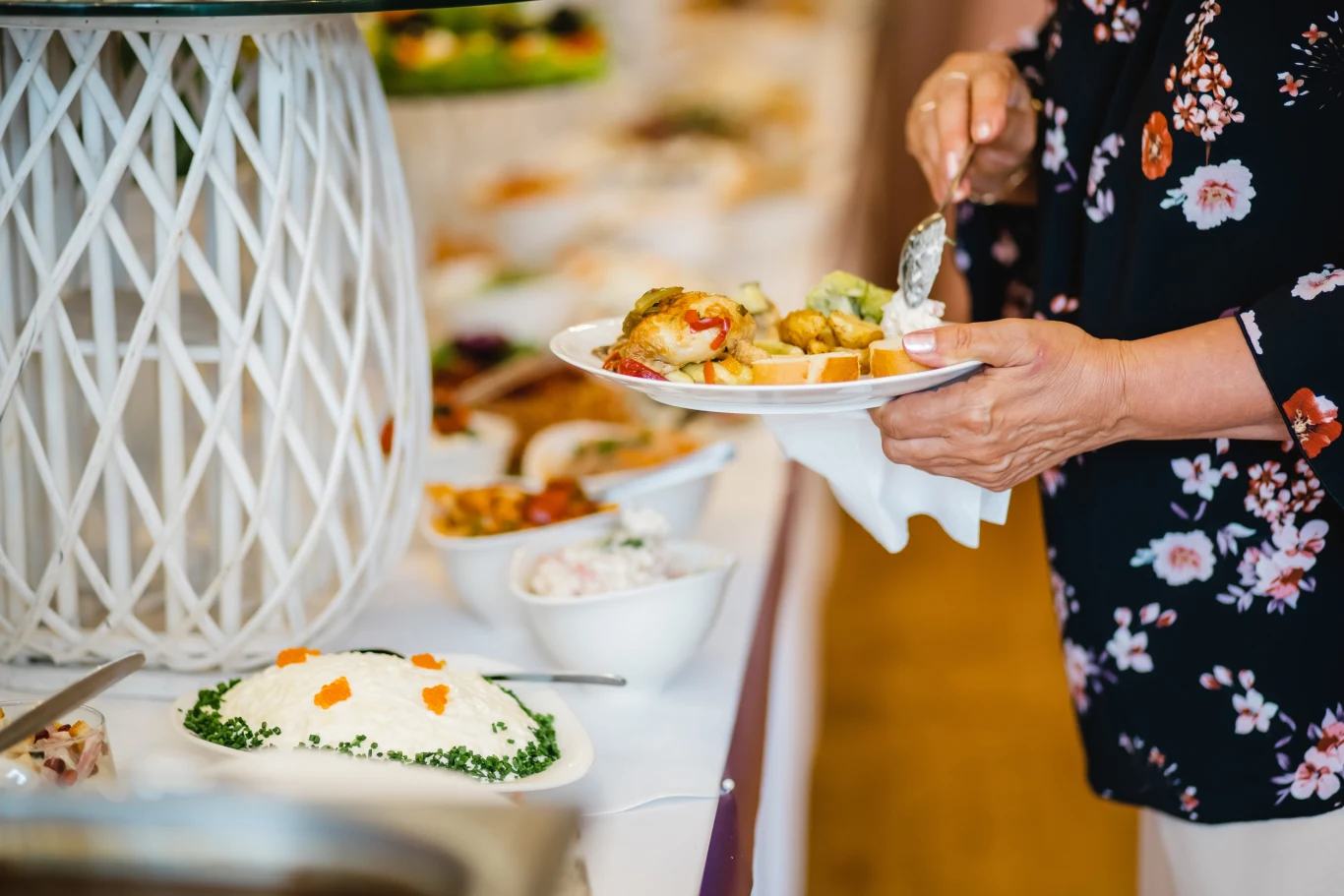 osoba w kwiecistej bluzce nakłada jedzenie na talerz przy bogato zastawionym stole z półmiskami pełnymi różnorodnych potraw