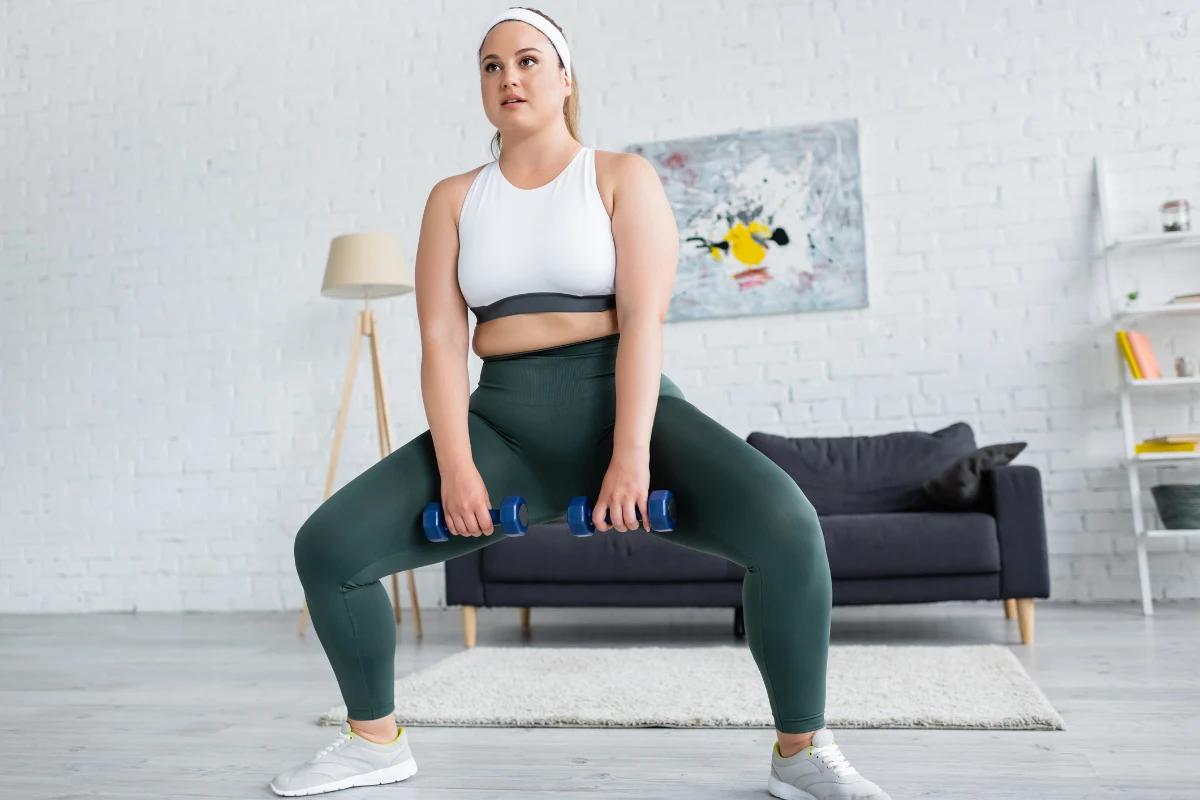 kobieta w sportowym stroju wykonuje przysiad sumo z hantlami w domowym salonie, skupiona na ćwiczeniu