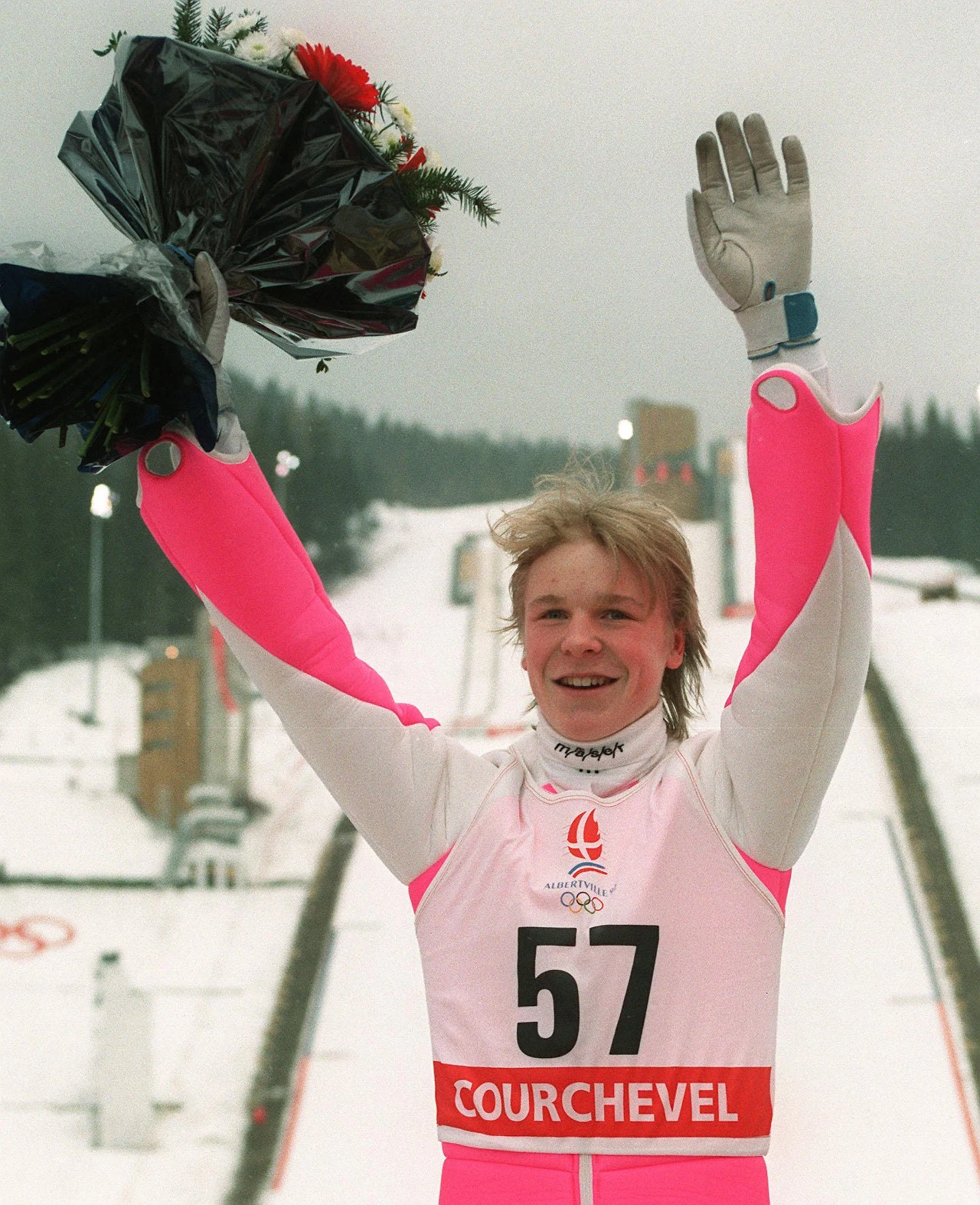 Skoczek narciarski w różowo-białym kombinezonie z numerem 57 stoi na tle skoczni, trzyma w górze bukiet kwiatów, uśmiecha się i macha do zgromadzonych osób, wokół zaśnieżone otoczenie i zimowa sceneria sportowa.
