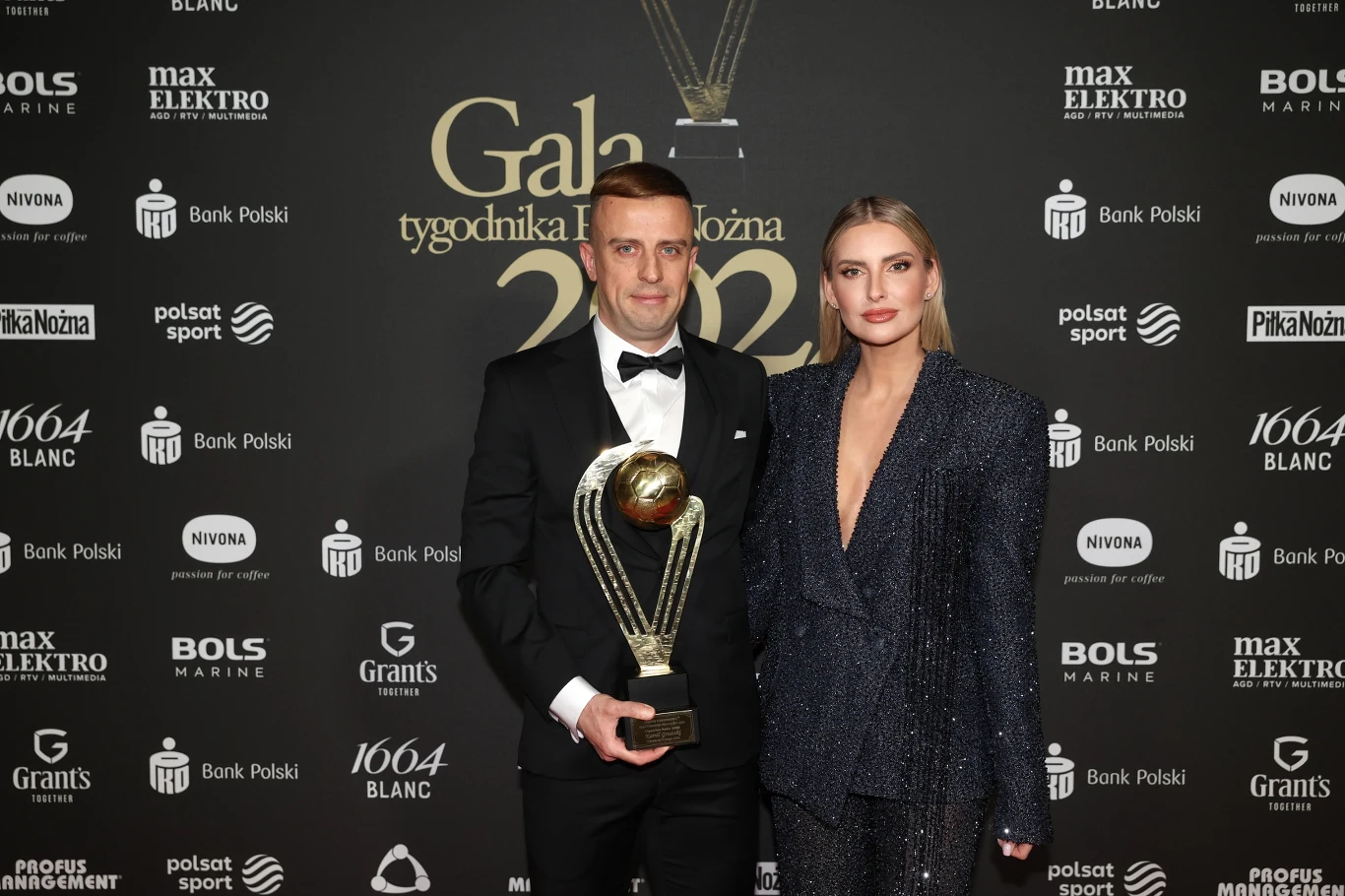 mężczyzna w smokingu trzymający dużą statuetkę nagrodową oraz kobieta w eleganckim, brokatowym garniturze na tle ściany z logotypami sponsorów podczas gali sportowej