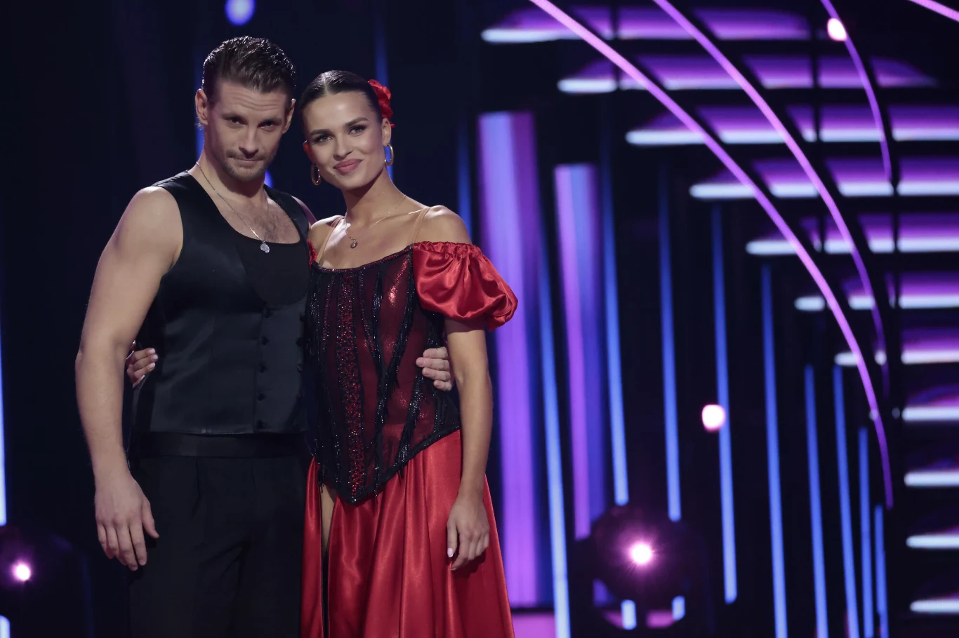 Tancerz w czarnej kamizelce i tancerka w eleganckiej czerwonej sukni pozują razem na scenie w otoczeniu fioletowych świateł oraz geometrycznych elementów scenografii.
