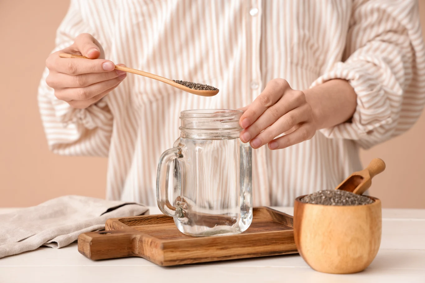 Osoba wsypuje nasiona chia z drewnianej łyżki do szklanki z wodą, obok stoi miseczka z większą ilością nasion oraz drewniana taca i ściereczka.