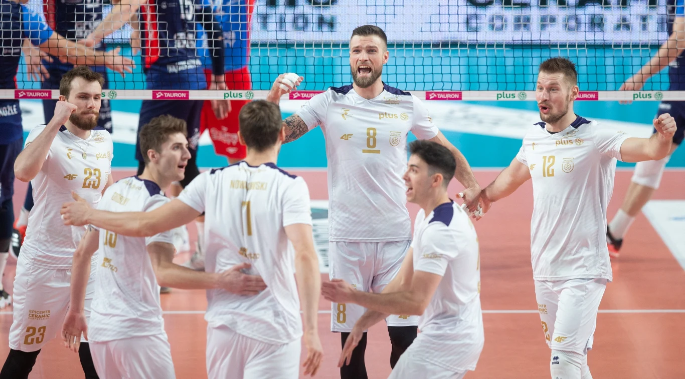 Sześciu siatkarzy w białych strojach świętuje zdobycie punktu na boisku, uniesione ręce i wyraz twarzy zawodników wskazują na radość i entuzjazm drużyny.