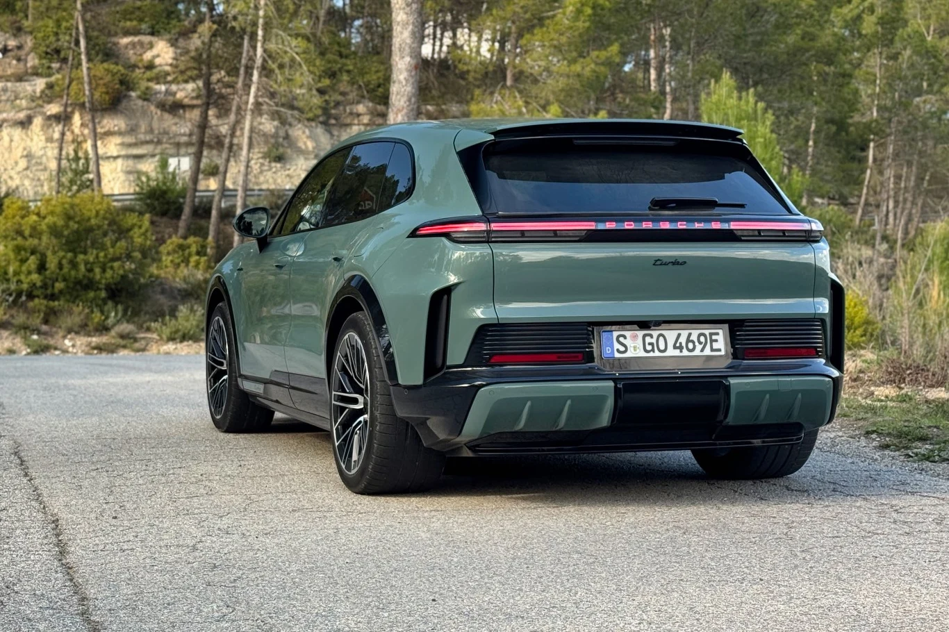 zielony SUV Porsche z napisem Turbo na tylnej klapie, charakterystycznym podświetlanym pasem LED i dużymi felgami, ustawiony na wąskiej asfaltowej drodze w otoczeniu drzew i skał