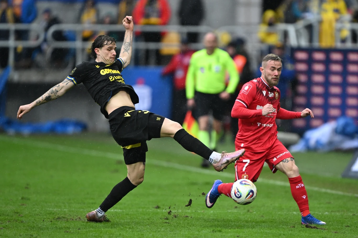 dwóch piłkarzy w dynamicznej walce o piłkę podczas meczu, jeden w czarnym stroju wykonuje wysoki wykop próbując przejąć futbolówkę, drugi w czerwonym stroju prowadzi piłkę przy nodze