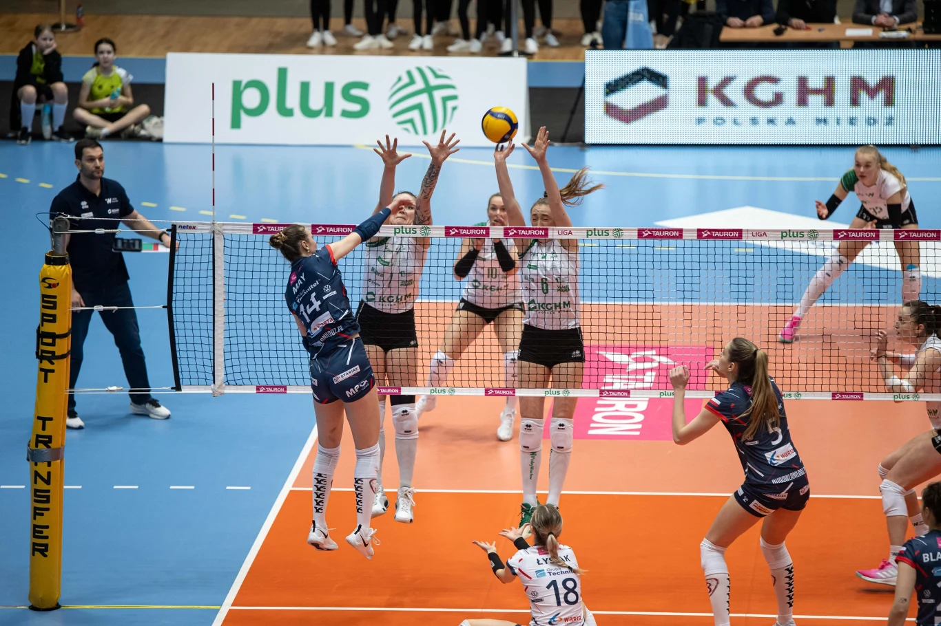 siatkarka drużyny w niebieskich strojach atakuje piłkę nad siatką, podczas gdy dwie zawodniczki drużyny w białych strojach próbują zablokować atak wyskakując jednocześnie do bloku, w tle widoczni pozostali członkowie drużyn i sędzia
