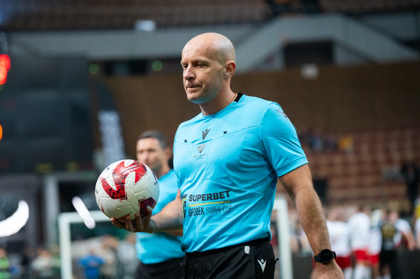 sędzia piłkarski w niebieskim stroju trzymający piłkę podczas meczu halowego, w tle widoczni inni uczestnicy spotkania oraz trybuny