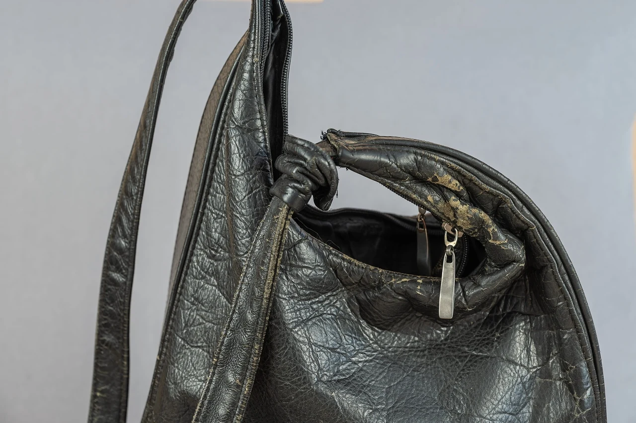 Zniszczona czarna skórzana torebka z widocznymi przetarciami i pęknięciami materiału, uchylonym zamkiem błyskawicznym oraz postrzępionymi brzegami w okolicy zamka.