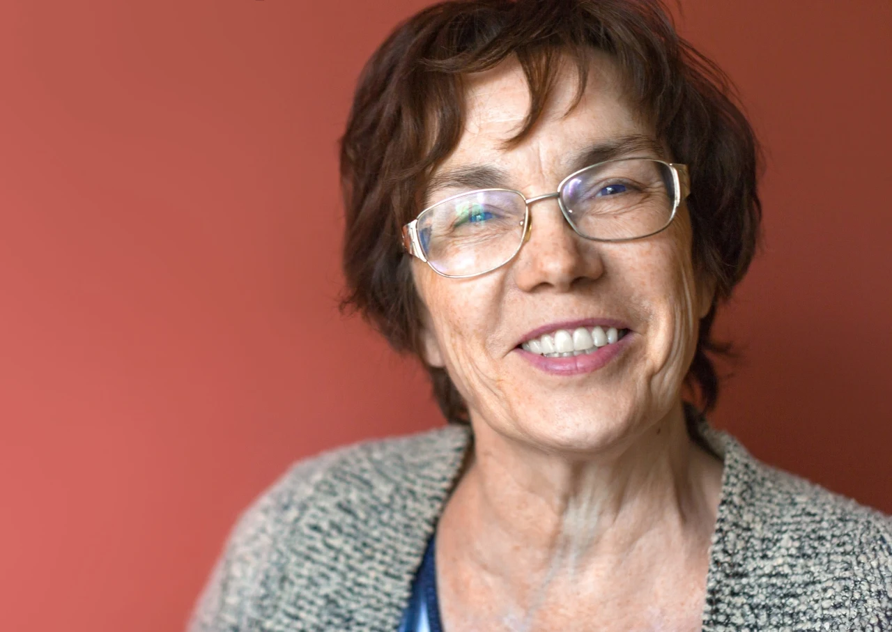 Uśmiechnięta starsza kobieta w okularach i swetrze na tle ciepłej, czerwonej ściany.