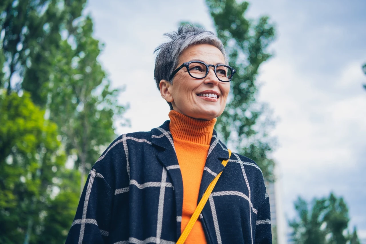 Uśmiechnięta kobieta z krótkimi, siwymi włosami i okularami ubrana w pomarańczowy golf oraz czarny płaszcz w kratę, stoi na tle zielonych drzew pod jasnym, lekko zachmurzonym niebem.