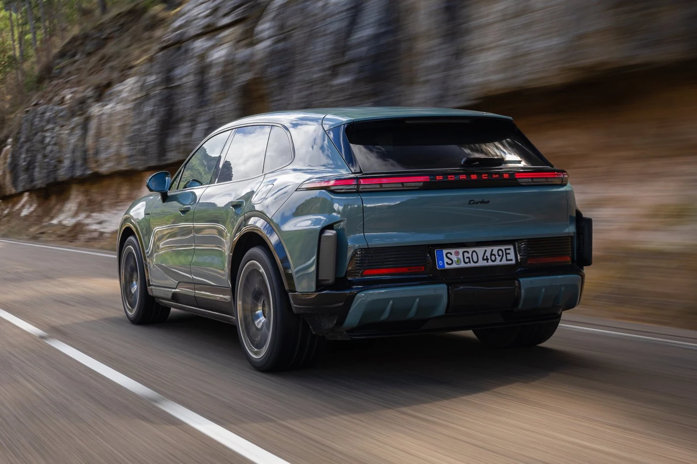 zielony samochód elektryczny typu SUV marki Porsche porusza się po asfaltowej drodze, otoczonej skałą i drzewami po lewej stronie, dynamiczny kadr podkreśla sportowy charakter pojazdu oraz nowoczesny design tylnej części z podłużnym pasem świateł LED