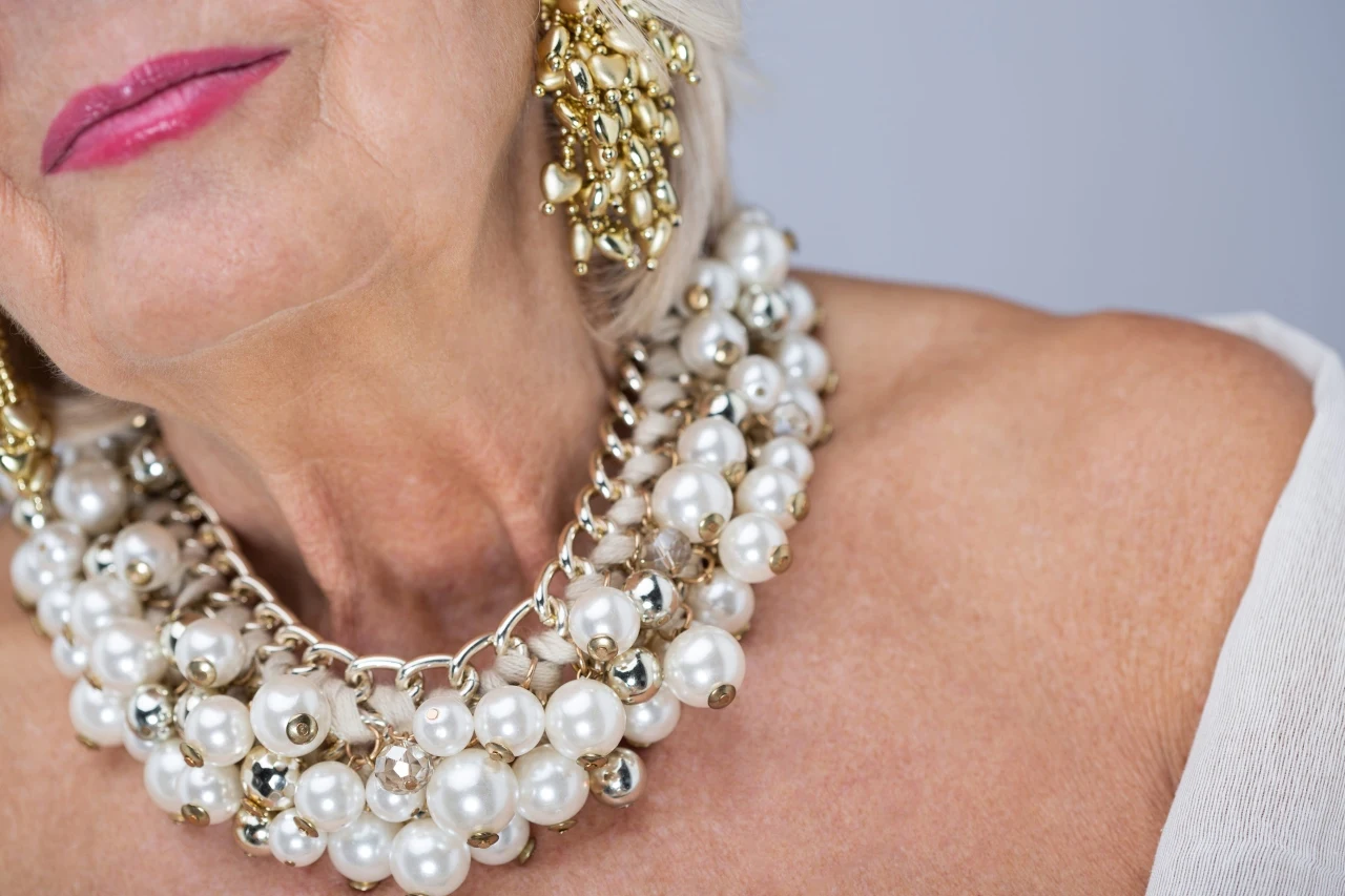 kobieta o jasnej cerze z wyrazistą różową pomadką na ustach, nosząca dużą, wielowarstwową kolię z białych i złotych pereł oraz efektowne złote kolczyki z ozdobnymi koralikami; jasne włosy i odsłonięte ramię dopełniają elegancki wizerunek