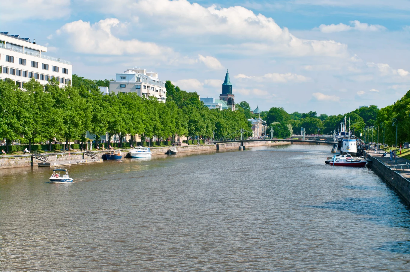 szeroka rzeka otoczona zielonymi drzewami i nowoczesnymi budynkami, kilka zacumowanych łodzi przy brzegu oraz łódź płynąca po wodzie, w tle wyłania się wieża zabytkowego kościoła