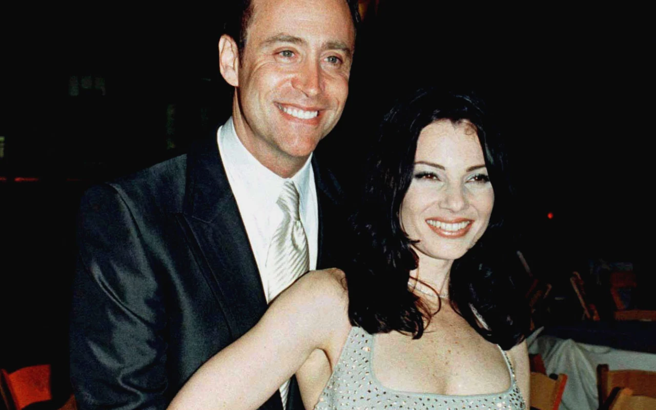 Uśmiechnięta para ubrana elegancko, mężczyzna w garniturze z jasnym krawatem obejmuje kobietę w srebrnej sukience na ramiączka, oboje patrzą w stronę obiektywu.