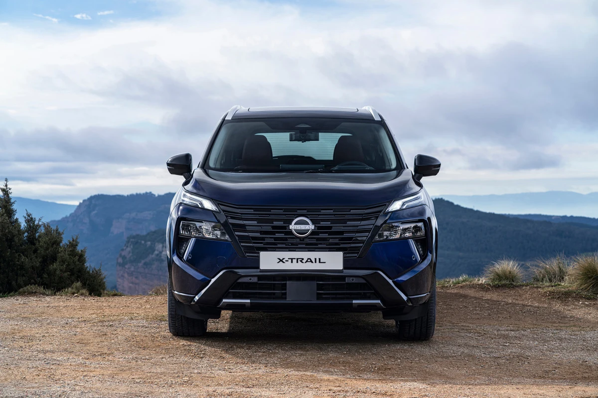 Granatowy SUV Nissan X-Trail stoi na szutrowej drodze, skierowany przodem do widza; w tle rozciąga się panorama gór z zachmurzonym niebem.