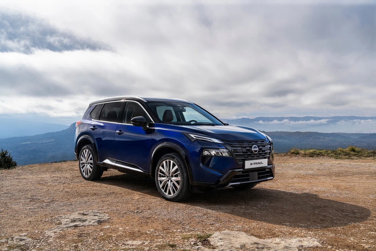 niebieski SUV Nissan X-Trail stoi na skalistym, górskim terenie pod pochmurnym niebem, z panoramicznym widokiem na odległe wzgórza i doliny