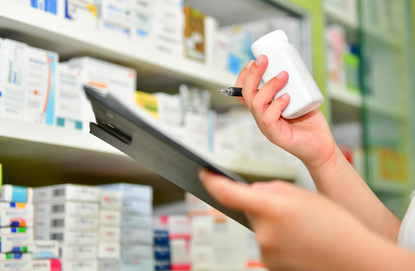 Utrudnienia w komunikacji z krajami Zatoki Perskiej utrudniają dostarczanie lekarstw, m.in. preparatów onkologicznych Na zdjęciu widoczne dwie dłonie, jedna trzyma podkładkę z kartkami, druga opakowanie leku. W tle półki z lekami