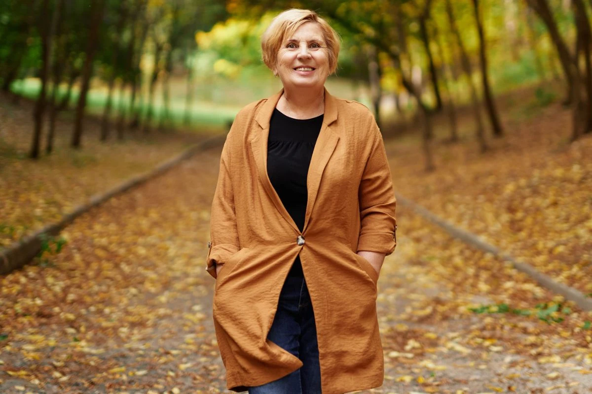 Uśmiechnięta kobieta w rudym płaszczu spaceruje jesienną aleją otoczoną drzewami z opadającymi żółtymi liśćmi.