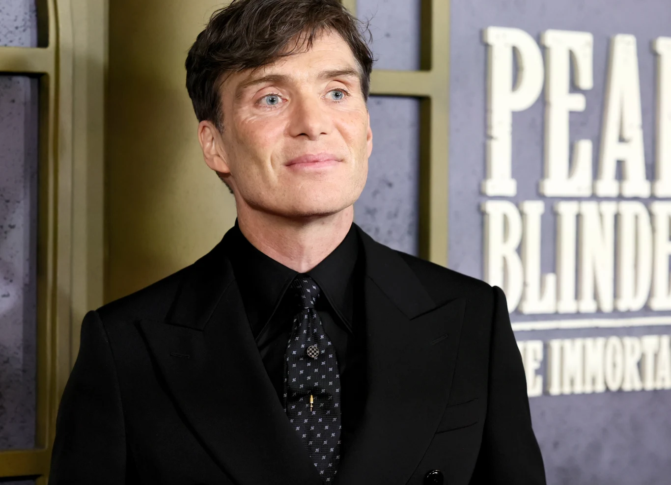 mężczyzna w eleganckim, czarnym garniturze i ciemnej koszuli z wzorzystym krawatem stoi na tle plakatu promującego serial Peaky Blinders, z wyraźnie widocznym tytułem z tyłu