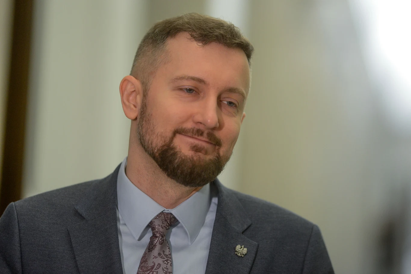 uśmiechnięty mężczyzna w garniturze z zakładaną marynarką i fioletowym krawatem, broda oraz krótko przycięte włosy, rozmyte jasne tło