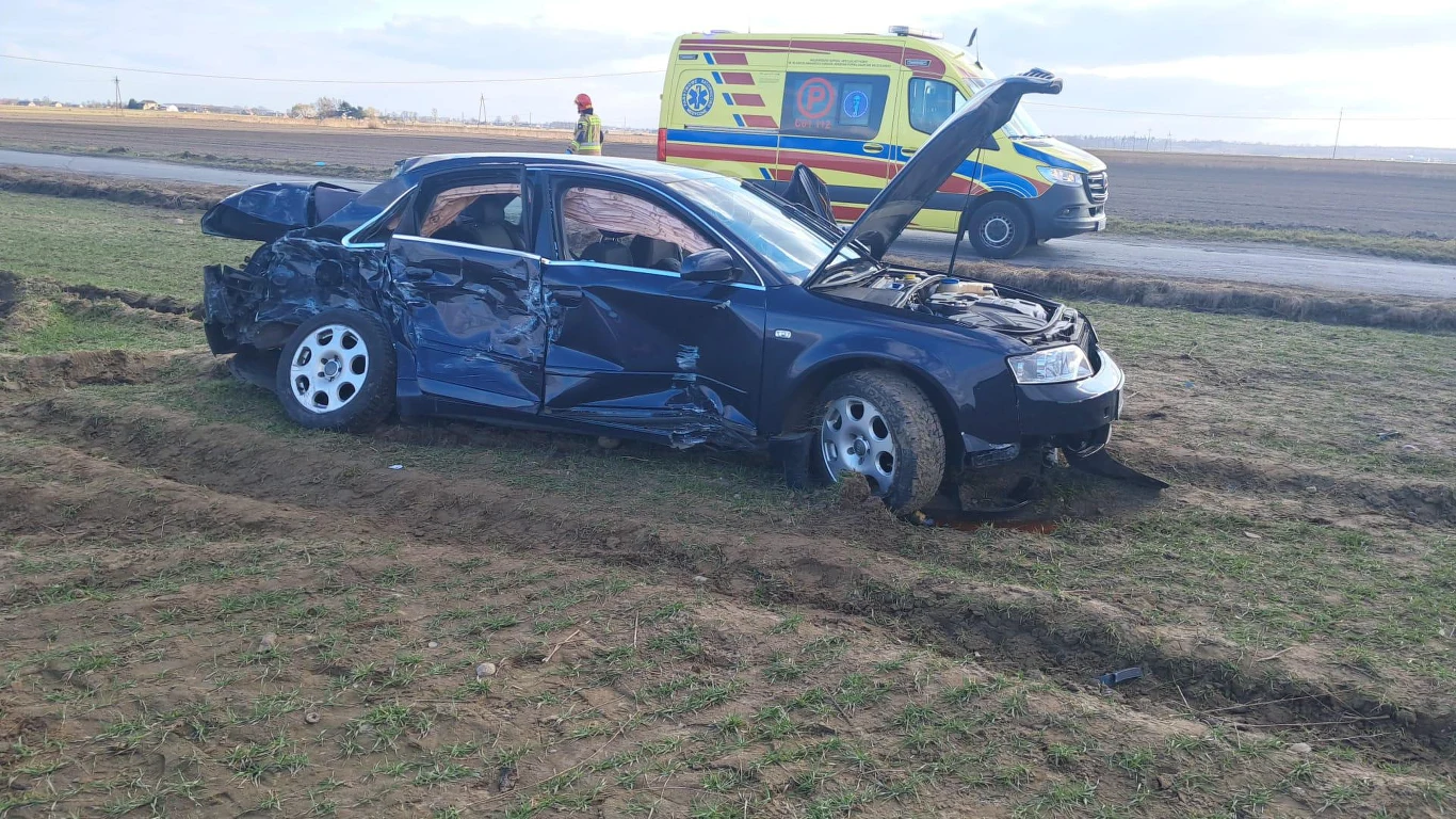 rozbity samochód osobowy z poważnie uszkodzonym bokiem i maską uniesioną do góry, w tle ambulans pogotowia oraz ratownik medyczny