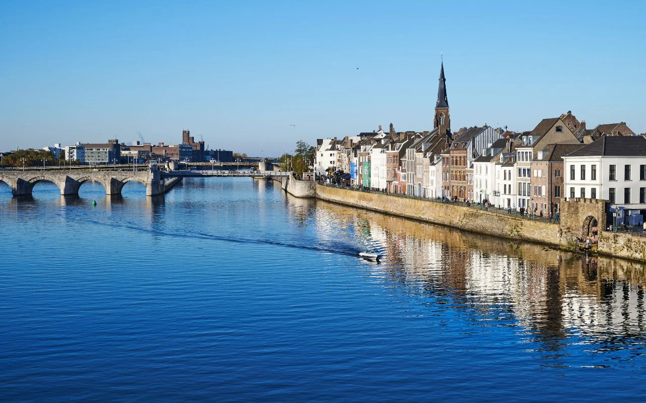 Zabytkowy kamienny most nad szeroką rzeką, łodzie na wodzie oraz rząd kolorowych kamienic z wysoką wieżą kościoła odbijających się w spokojnej tafli wody.