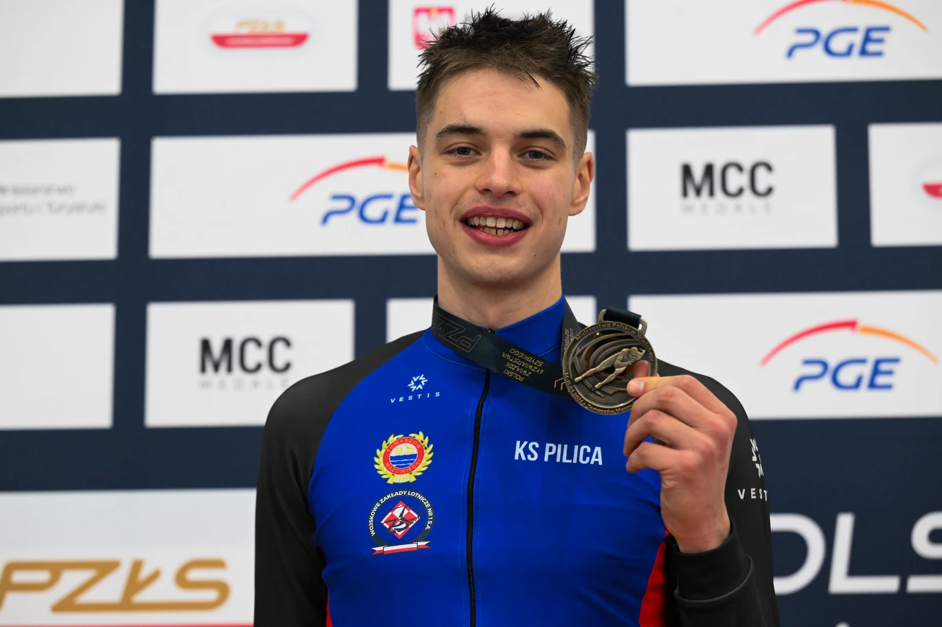 młody sportowiec w niebiesko-czarnym stroju kolarskim z wyraźnym napisem KS Pilica, uśmiecha się i prezentuje medal zawieszony na czarnej wstążce, w tle widoczne są logotypy sponsorów oraz tablica z nazwami partnerów wydarzenia sportowego