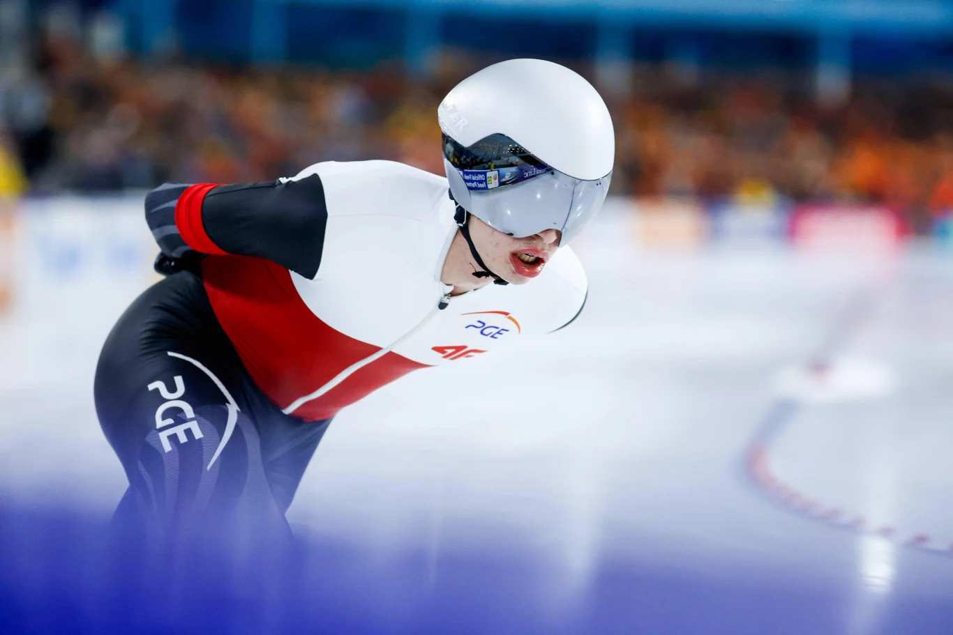 łyżwiarz szybki w kombinezonie startowym z polskim akcentem kolorystycznym, w aerodynamicznym kasku i okularach, pochylony do przodu podczas wyścigu na torze lodowym